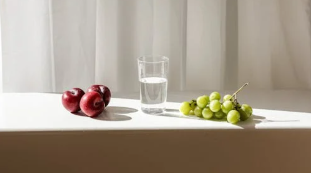 Glas vand, røde æbler og grønne druer på vindueskarm